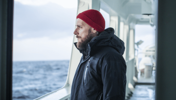 Mann på dekk, står langs rekka å ser utover havet og horisonten. Kled i svart jakke og raud lue